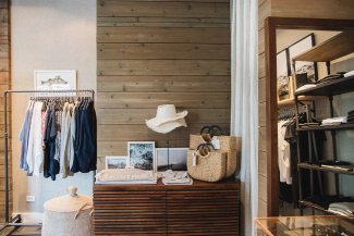 Clothing boutique with shirts on a rack, straw hats, and woven bags displayed on wooden shelves. Cozy, minimalist interior design.