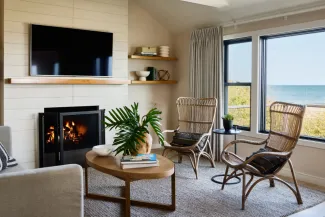 Cozy living room with a fireplace, two wicker chairs, a wooden coffee table, and a view of the beach through large windows.