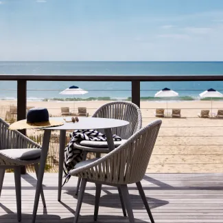 Two chairs and a table on a deck overlooking a sandy beach with white umbrellas and the ocean in the background.