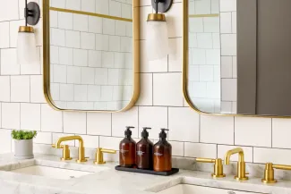 Elegant bathroom with two gold faucets, a white tiled wall, two mirrors, and three brown soap dispensers on a marble countertop.