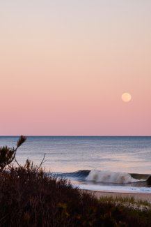 View from the beach of the moon on the horizon over the ocean at dusk.