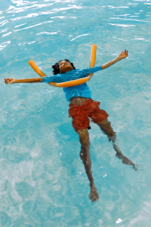 A child floats in blue water with an orange pool noodle