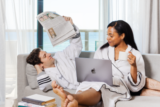 A couple in robes read the newspaper and browse on a laptop with the ocean in the background