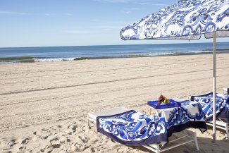 Beach with patterned blue loungers and umbrella on white sand, facing the calm sea under a clear sky.