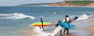 Surfers on a sandy beach carrying colorful surfboards toward the ocean, with waves and rocky cliffs in the background.