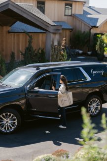 A person with a tote bag is entering a black SUV parked outside a modern wooden house on a sunny day.