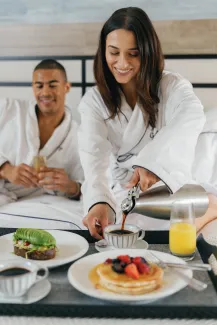 Couple in bathrobes enjoying breakfast in bed. Woman pours coffee next to pancakes and fruit, avocado toast, and orange juice.