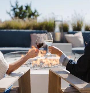 Two people clink wine glasses while sitting in chairs by a fire pit outdoors.