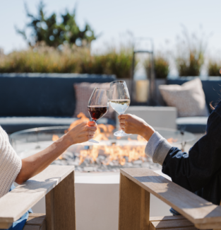 Two people clink wine glasses while sitting in chairs by a fire pit outdoors.