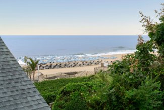 Ocean view with sandy beach, rows of umbrellas, and palm trees, framed by a rooftop and lush greenery under a clear blue sky.