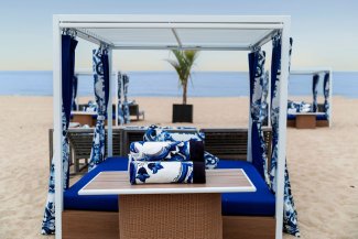 Beach cabana with blue and white decor on sandy shore, calm sea and sky in background, single palm tree visible in distance.