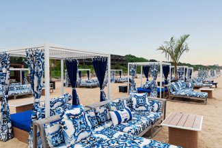 Beachside cabanas with blue patterned cushions and curtains, set on sandy shore with a clear sky and a solitary palm tree.
