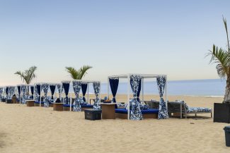 Beach cabanas with blue and white drapes line the sandy shore, facing a calm sea under a clear sky. Sparse palm trees are scattered around.