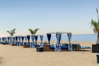Cabanas with blue and white curtains on a sandy beach, palm trees nearby, calm sea under a clear sky.