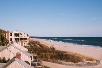 Coastal homes line a sandy beach under a clear blue sky, with gentle ocean waves in the distance.