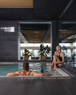 Indoor pool setting with a man swimming and a woman sitting on the edge. Large windows in the background, plants visible. "3 Feet 6 Inch Deep" marked on the pool edge.