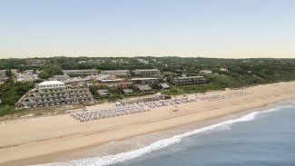 Aerial view of a beach with rows of umbrellas, buildings set back among green trees, and gentle waves on the sandy shore under a clear sky.