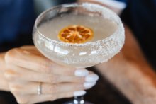 Close up of woman holding cocktail in fine glassware with citrus garnish and sugar-lined rim.