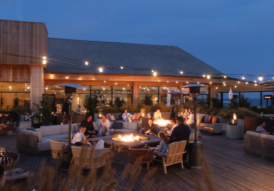 Outdoor restaurant patio at dusk with people gathered around glowing fire pits, surrounded by string lights and modern seating.